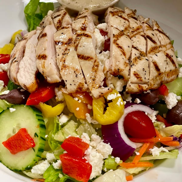 Grilled chicken salad with feta, olives, bell peppers, cucumbers, and a side of dressing.