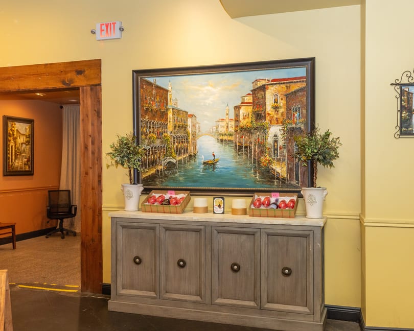 Restaurant interior with a large Venice canal painting above a cabinet displaying decorative items.