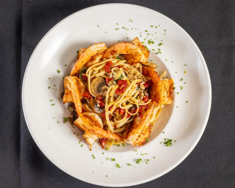 Linguine pasta with grilled shrimp, mushrooms, sun-dried tomatoes, and parsley in a white bowl.