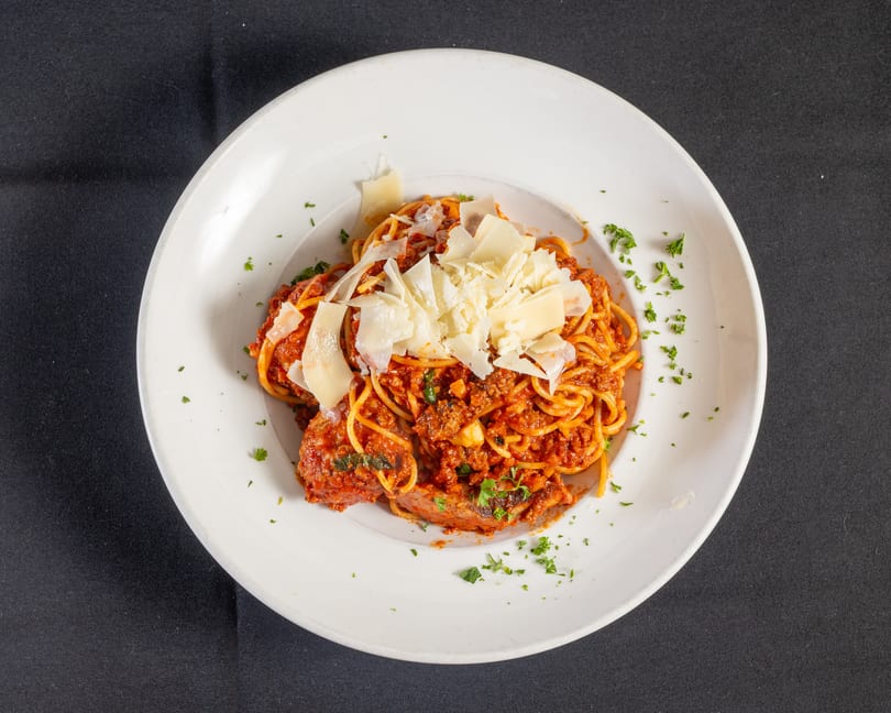 Spaghetti and meatballs with rich meat sauce, shaved Parmesan, and fresh parsley in a white bowl.