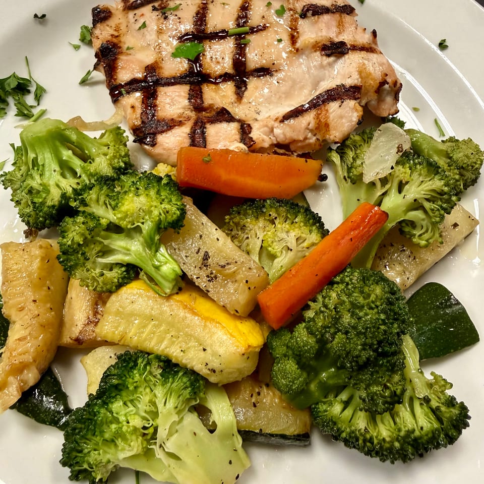 Grilled chicken breast with char marks, served alongside steamed broccoli, carrots, and squash on a white plate.