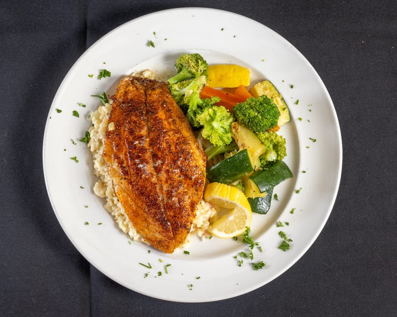 Pan-seared fish with creamy rice, colorful steamed vegetables, and a lemon wedge.