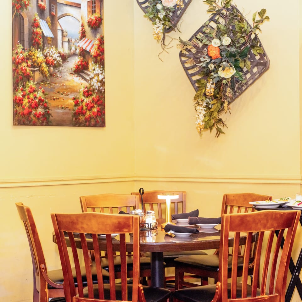 Cozy restaurant corner with a set table, wooden chairs, a vibrant painting, and decorative floral baskets.