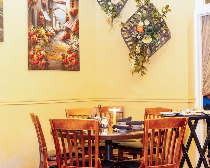 Cozy restaurant corner with a set table, wooden chairs, a vibrant painting, and decorative floral baskets.